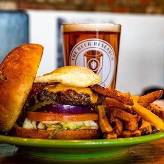 A cheeseburger topped with a fried egg on a plate with fries sits on a table beside a glass of beer.